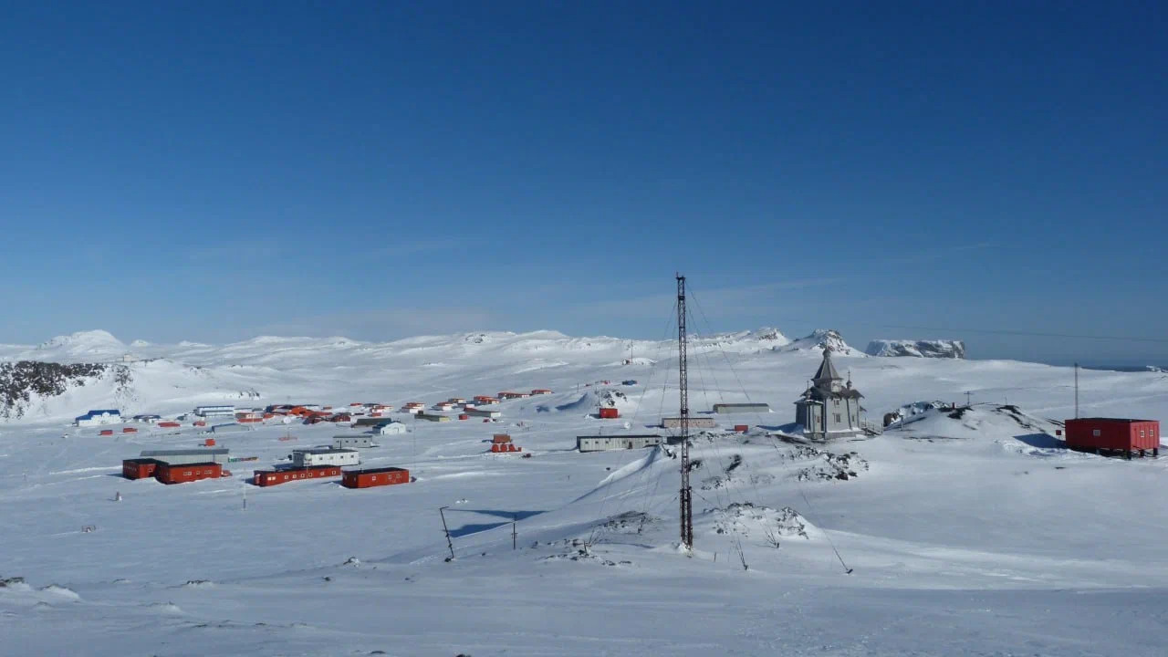 Antarctic research station with red and white buildings, including a church, set in a snowy landscape under a clear blue sky.

