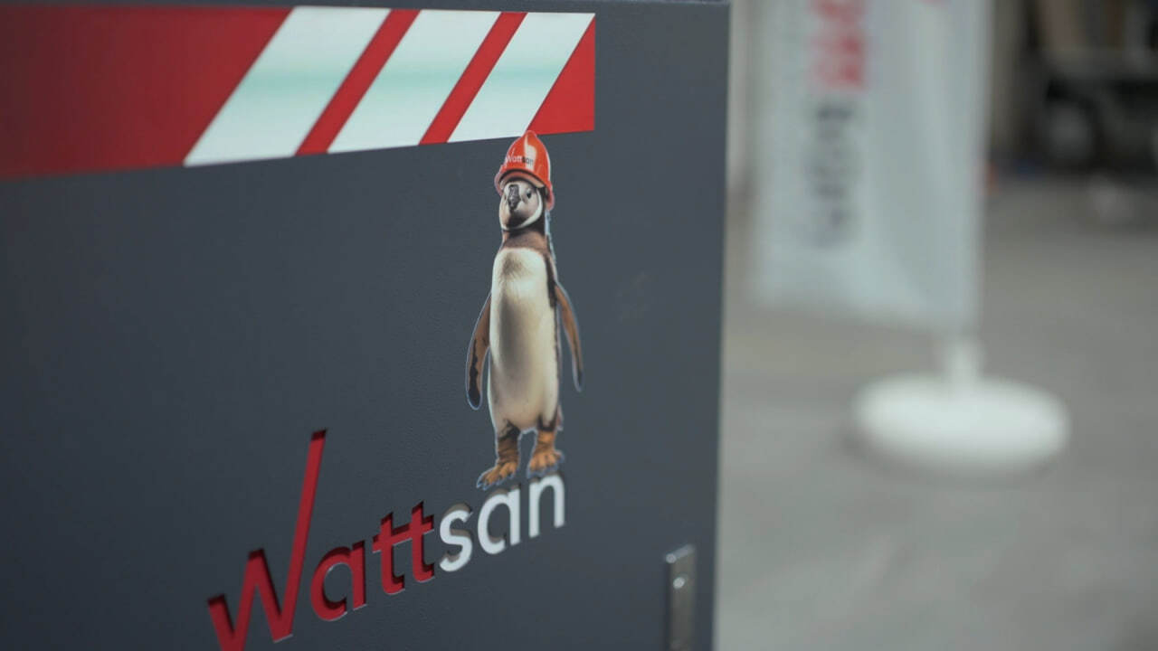 Penguin sticker wearing a red hard hat labeled “Wattsan” on a dark panel, with partial branding visible in the background.
