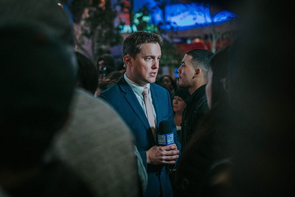 News reporter conducting an interview in a crowded outdoor setting at night.