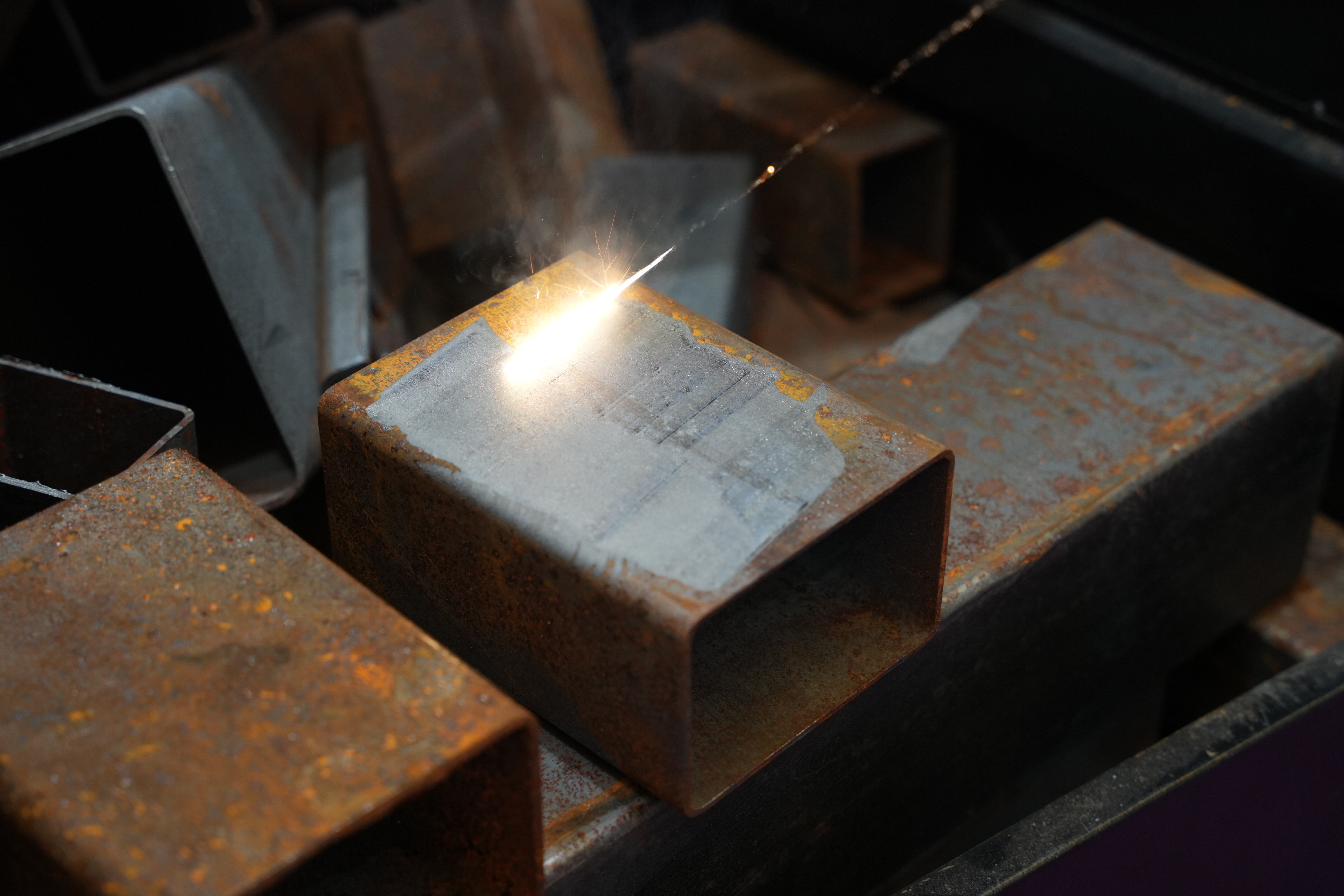 Worker using a bright welding torch on rectangular metal tubes, with sparks flying during the cutting process.
