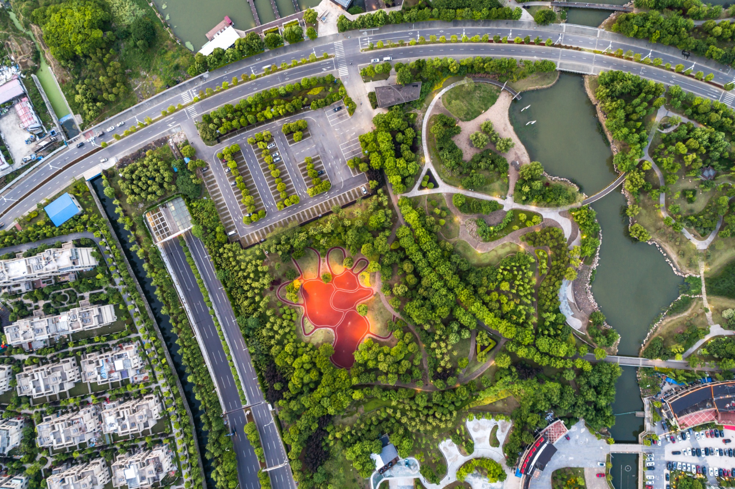 Aerial view of an urban park with winding paths, green trees, and a red, organically shaped playground surrounded by roads and residential buildings.