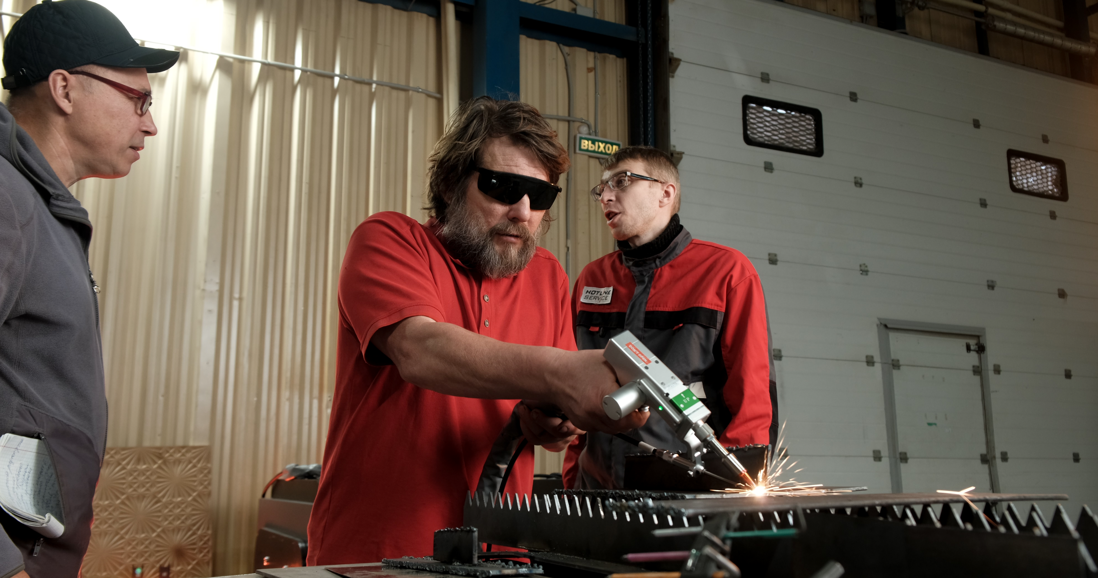 Worker in a protective mask cuts metal with a handheld tool in a workshop, while two others observe amid industrial equipment.
