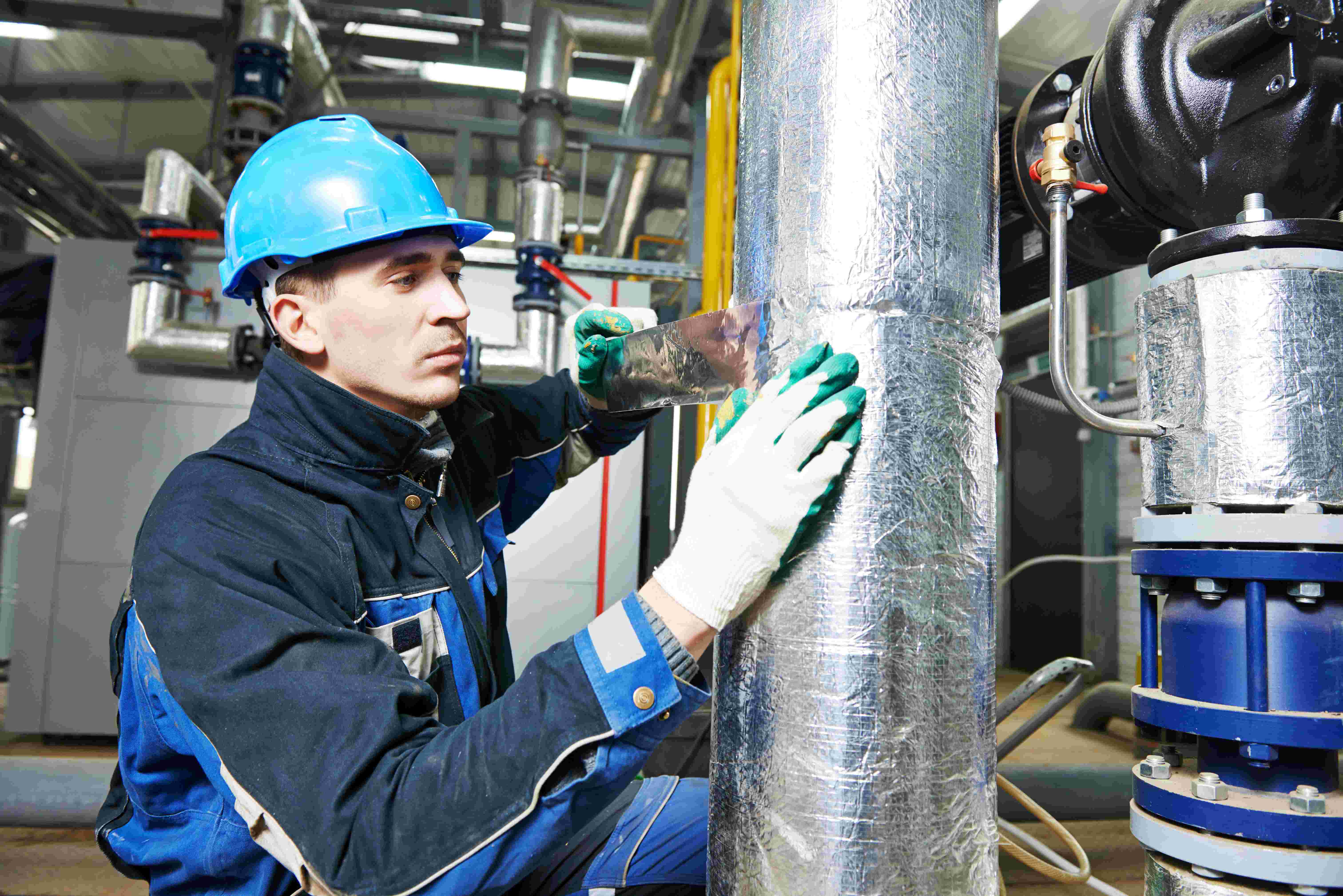 Worker in blue protective clothing secures insulation around a large industrial pipe using reflective tape.