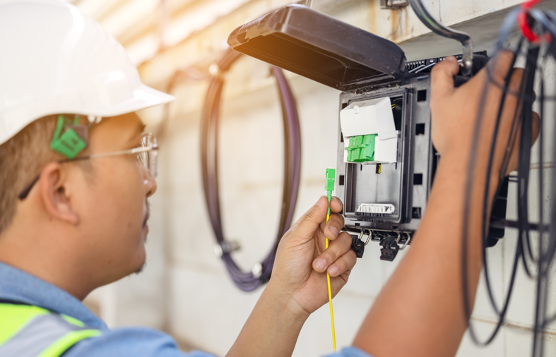 Technician in a hard hat and safety vest connects a green fiber optic cable to a wall-mounted terminal box.