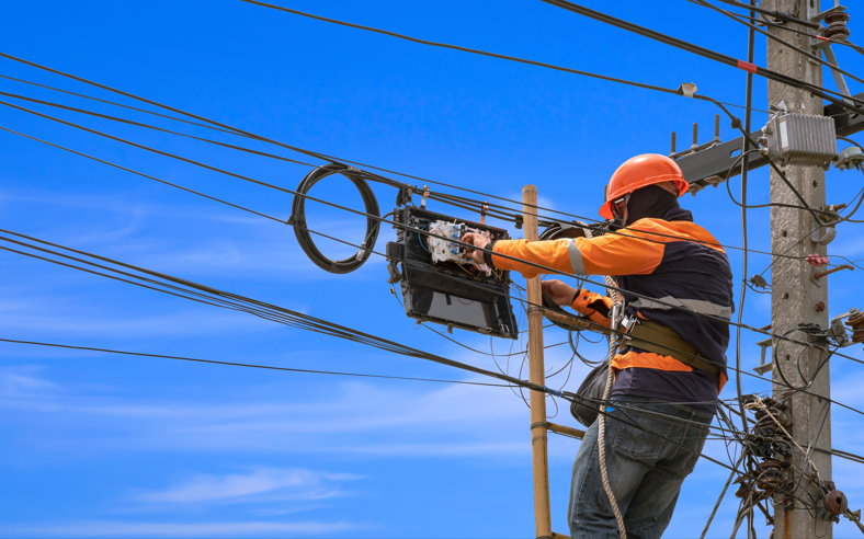 Worker in helmet and high-visibility vest services equipment on a utility pole, surrounded by electrical wires against a clear blue sky.