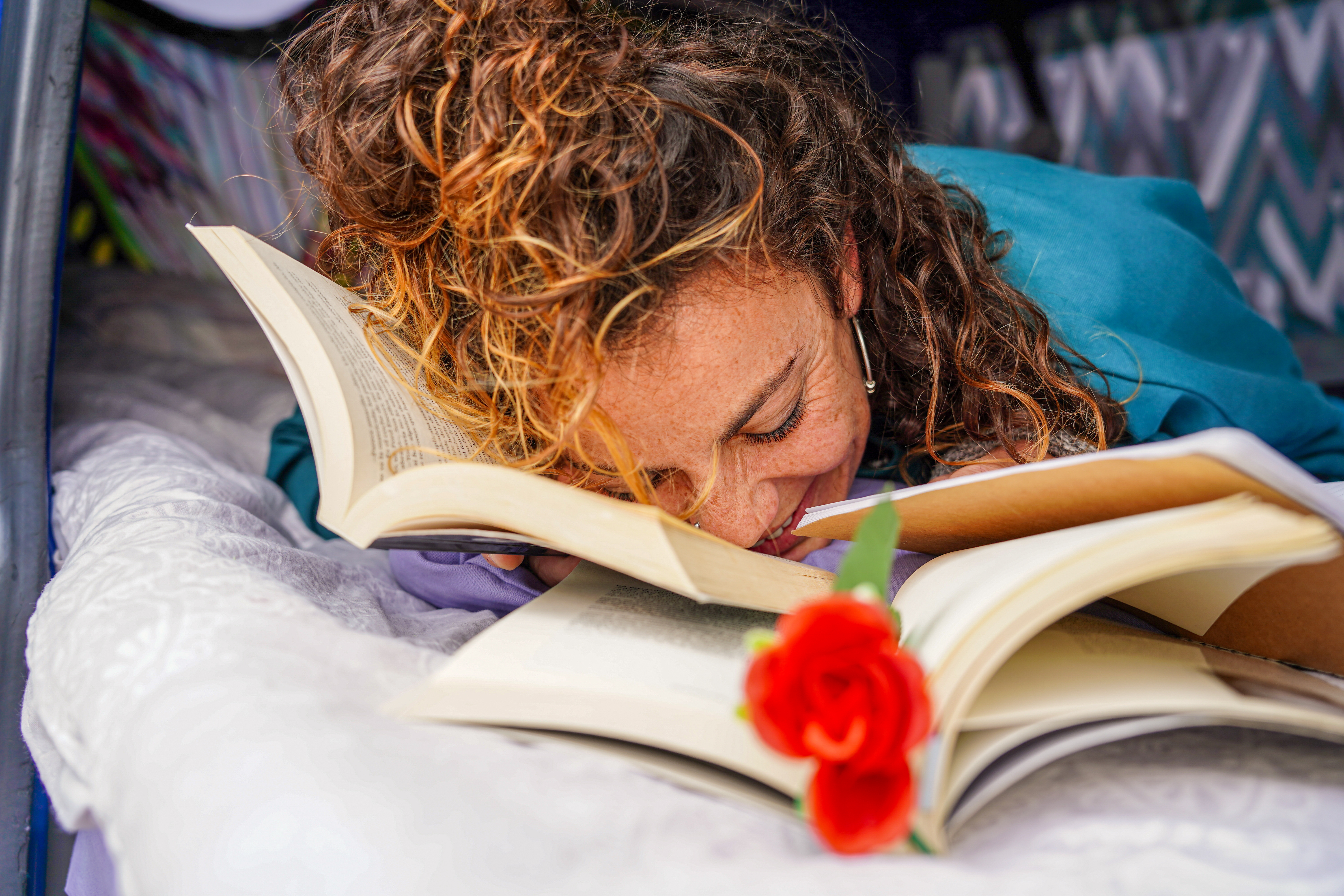Person with curly hair lies on a bed surrounded by open books, with a red rose resting on one of them.