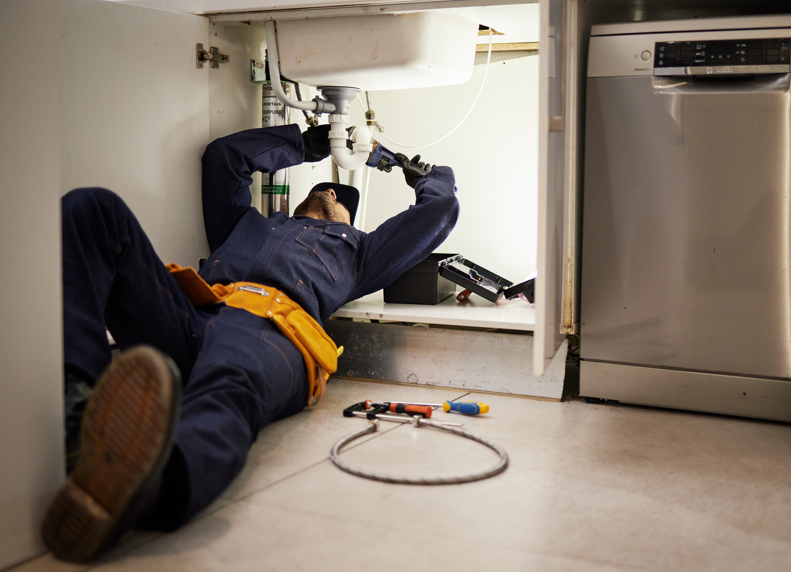 A plumber lying on the floor under a kitchen sink, using tools to repair the plumbing system.