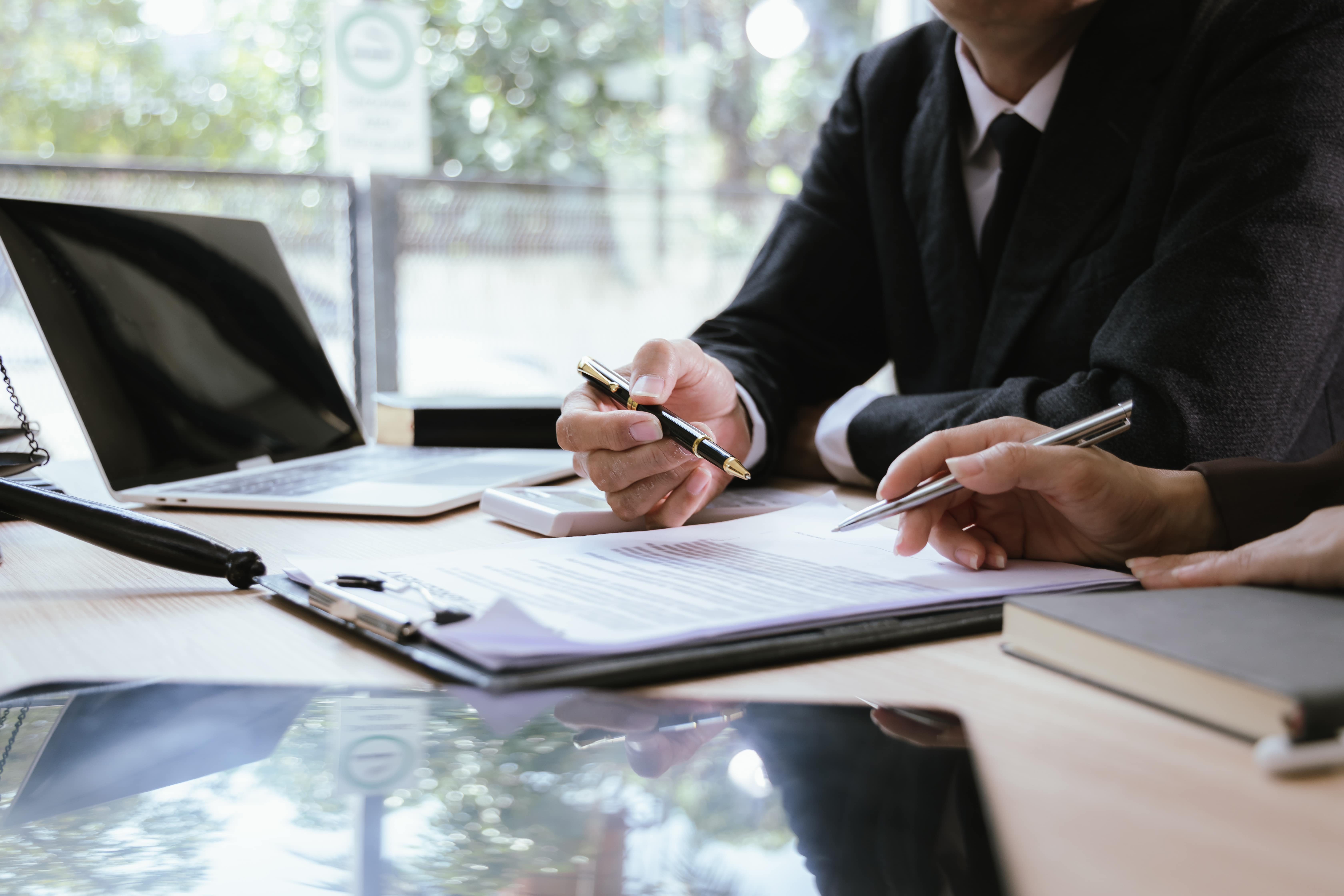Close-up of two business professionals in suits reviewing and discussing documents at a desk, with a laptop, clipboard, and other office supplies around them.