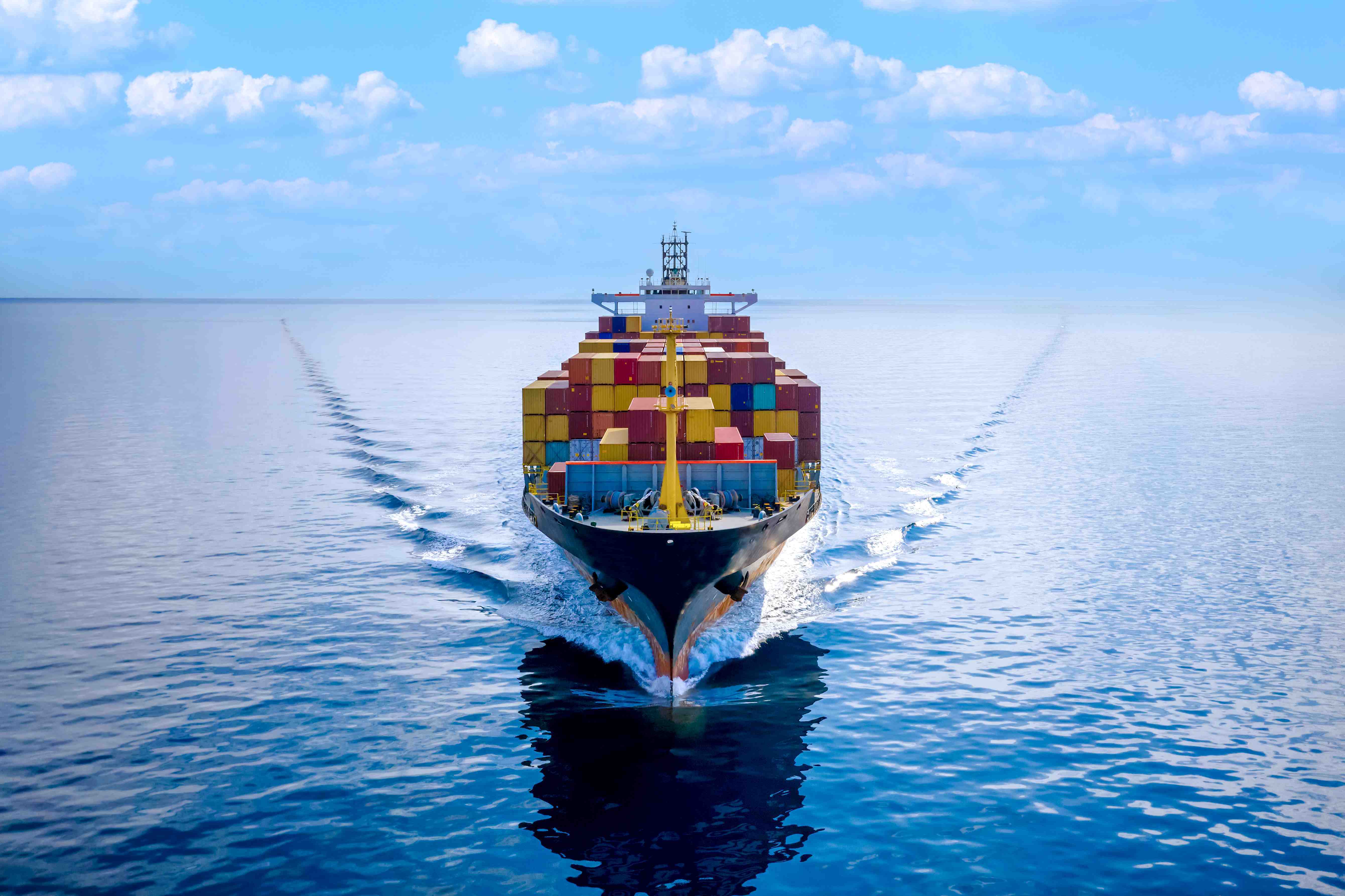 A large cargo ship filled with shipping containers sailing on the open sea under a cloudy sky.