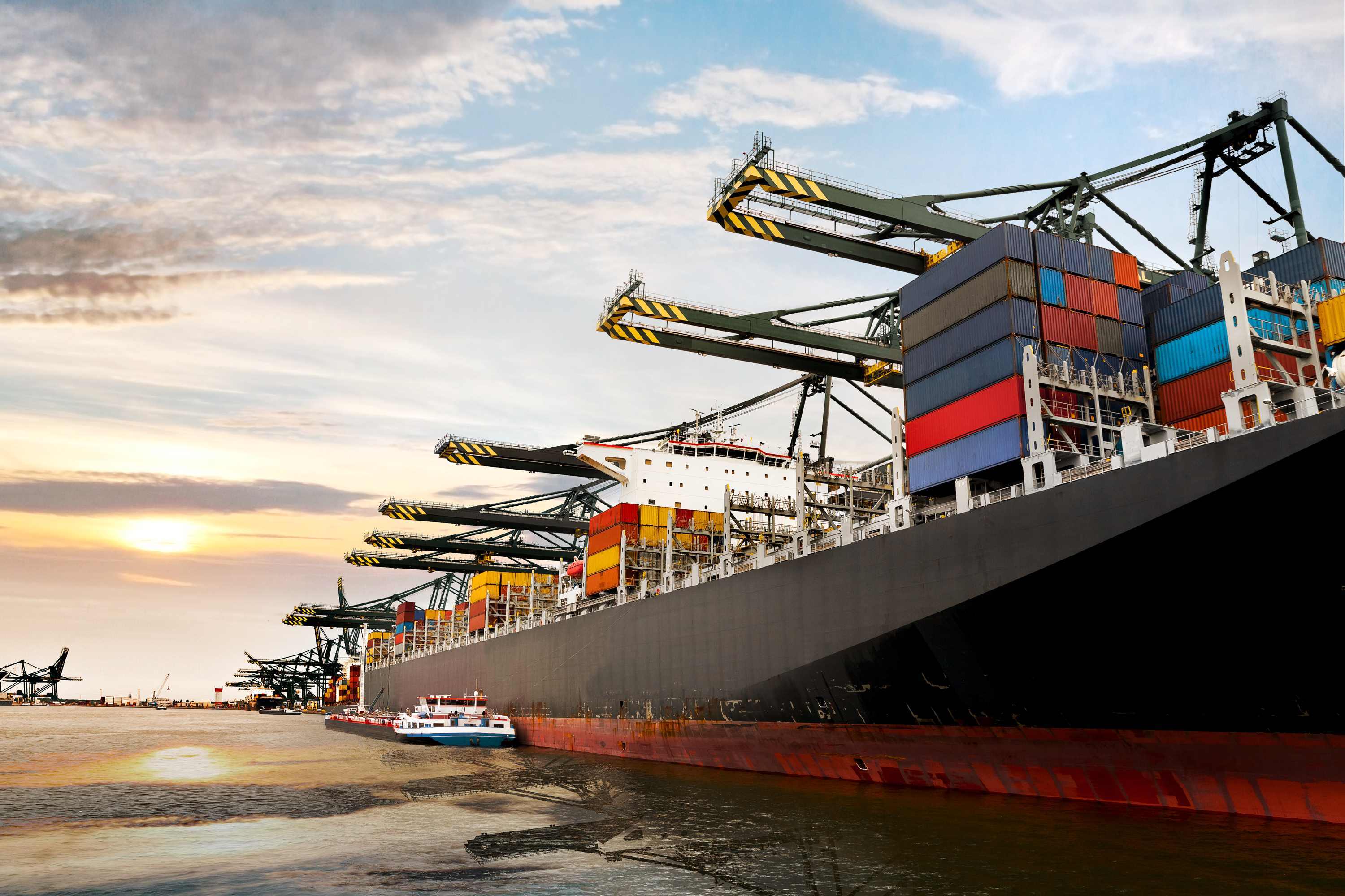 A large container ship with colorful shipping containers docked at a port with cranes and industrial buildings.