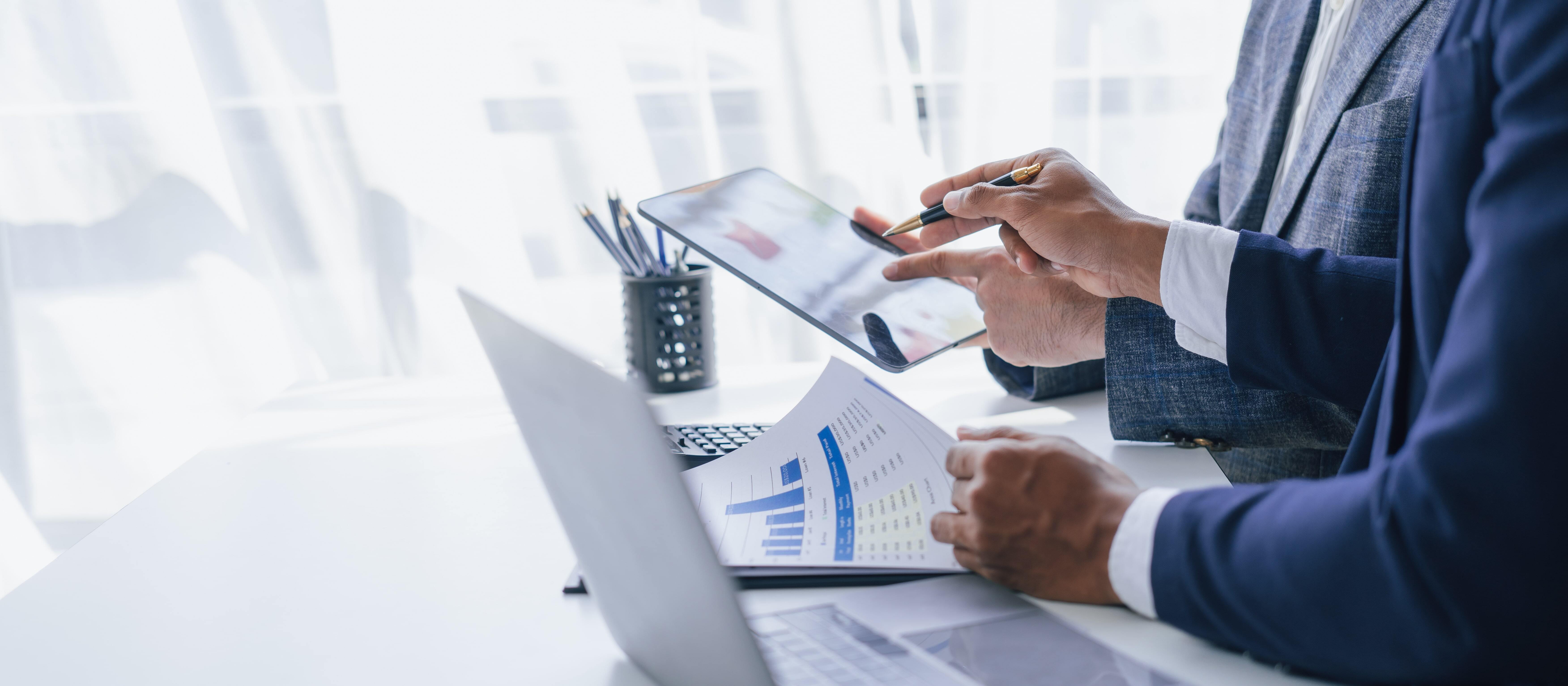 Two professionals in business attire collaborate at a desk, reviewing documents and a tablet displaying charts.