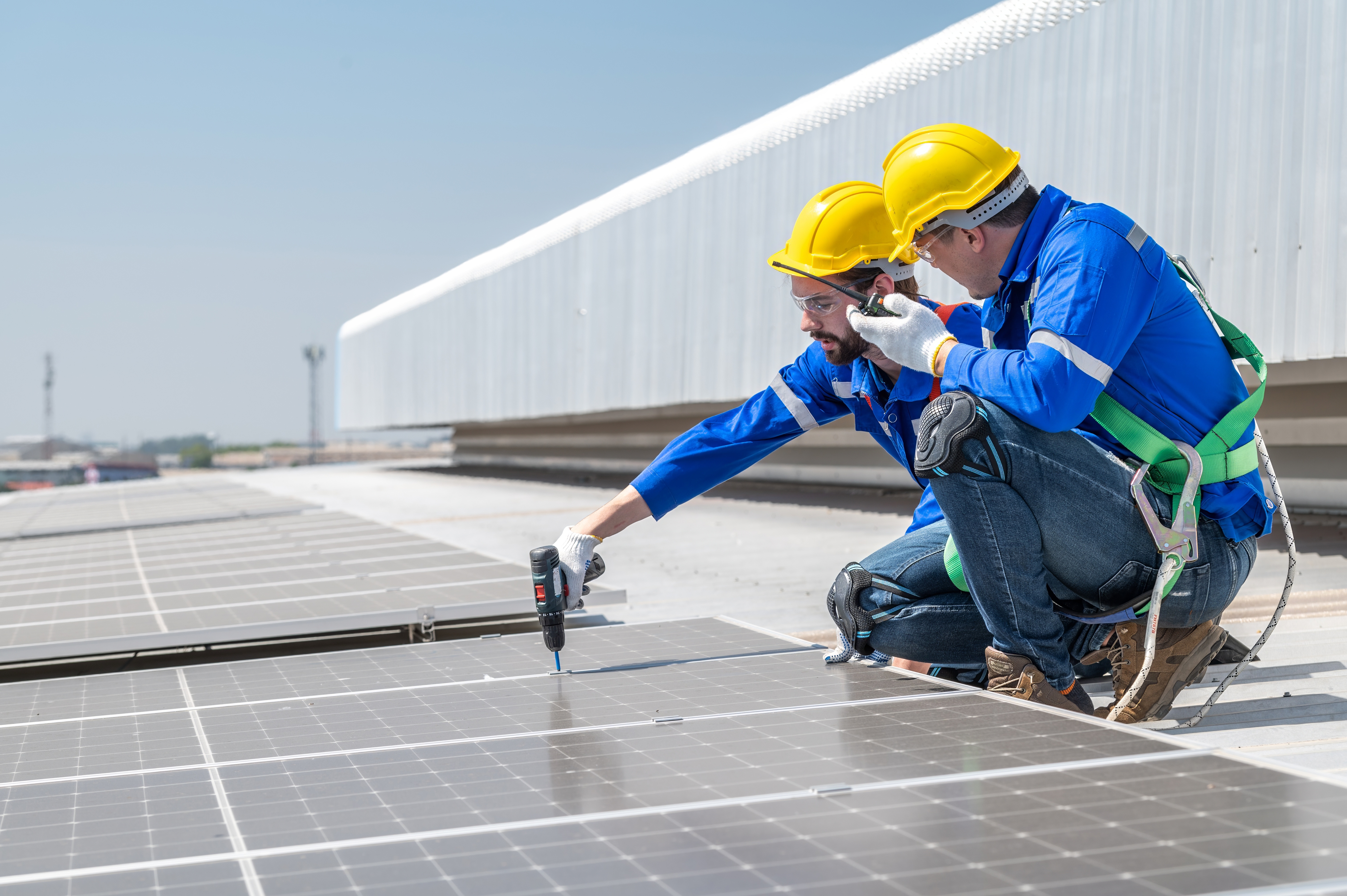 Two workers in safety gear installing solar panels on a rooftop, using tools and pointing at the panel surface.