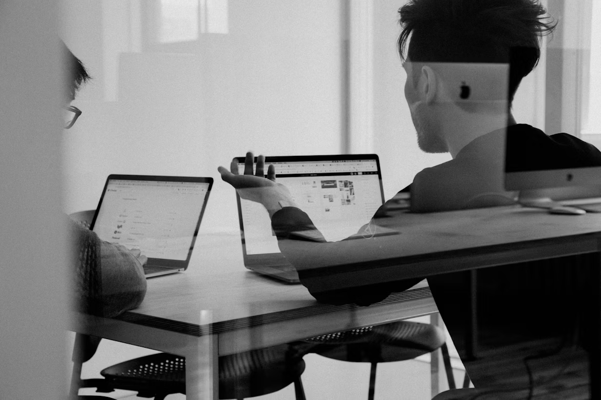 A black and white photo shows two people collaborating at a desk with laptops, seen through a glass reflection.
