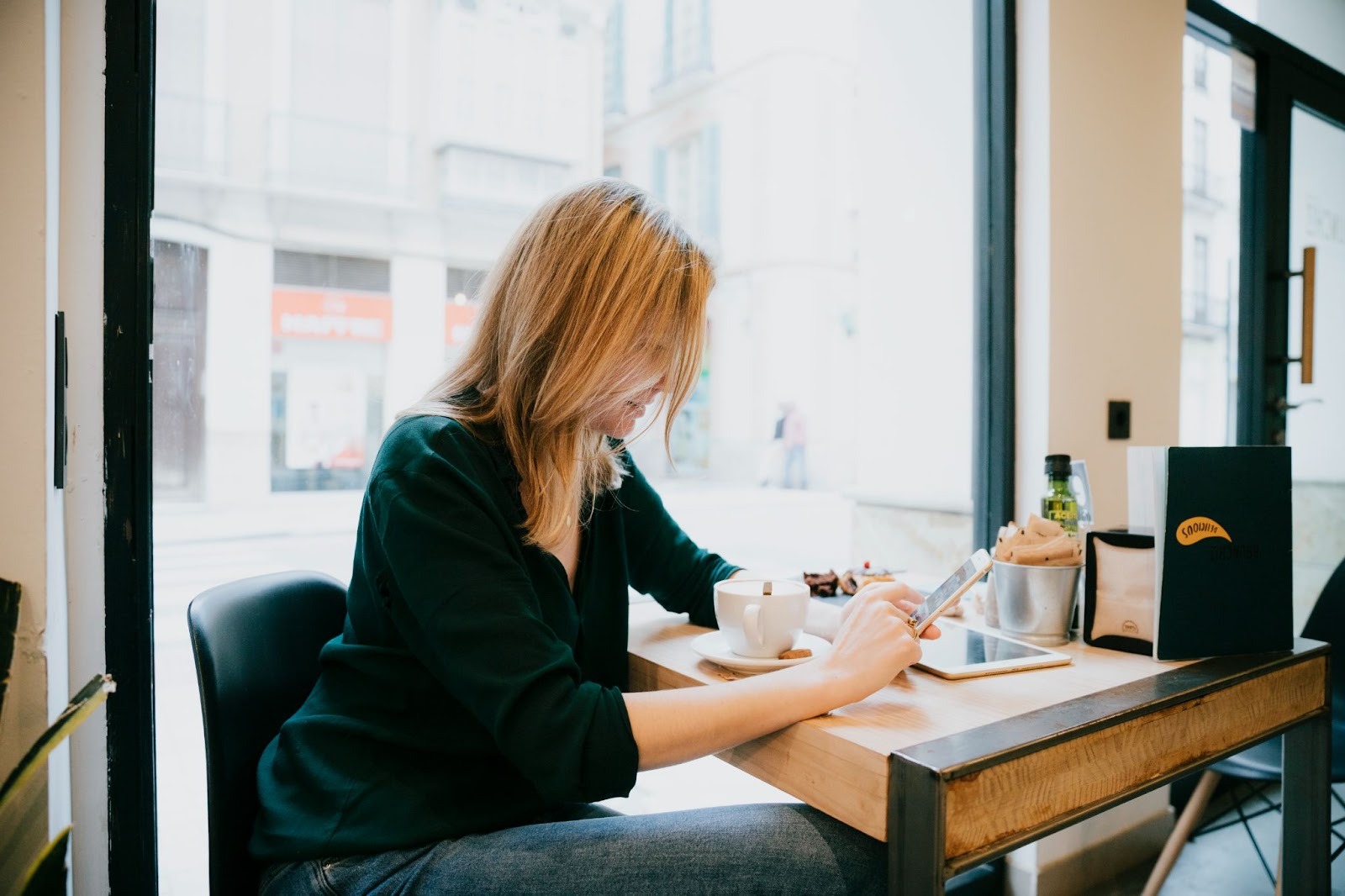 A blonde woman in a black blazer using her smartphone at a cafe table with a cup of coffee and a tablet.