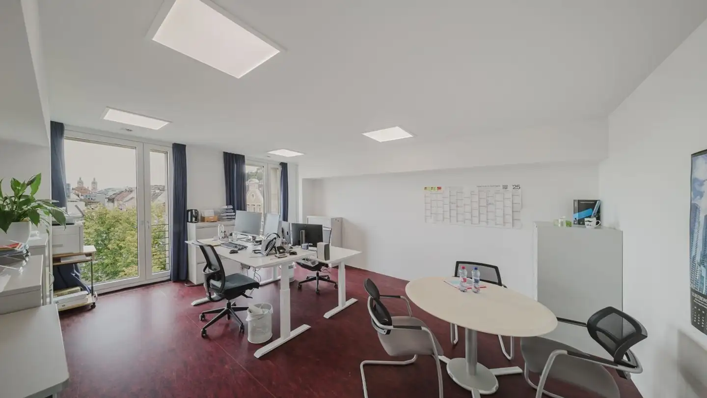 A bright, minimalist office featuring white height-adjustable desks, ergonomic chairs, and a round meeting table with large windows overlooking a city.