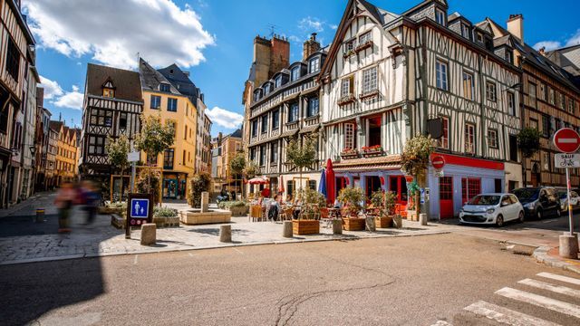 A charming European street scene featuring historic half-timbered buildings, an outdoor cafe with red umbrellas, and a cobblestone plaza under a bright blue sky.