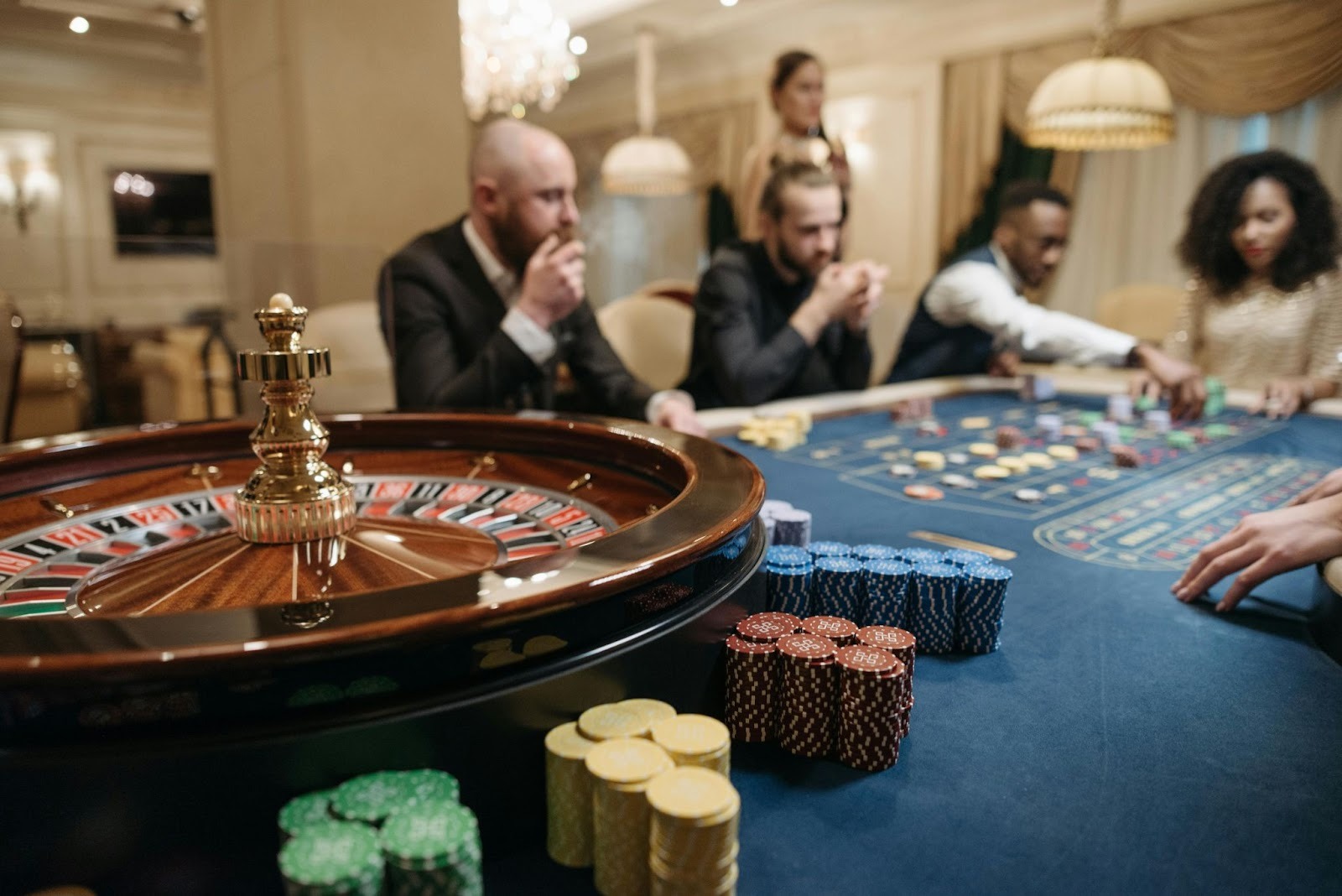 A close-up view of a spinning roulette wheel in the foreground with people playing at a blue betting table in a brightly lit casino.