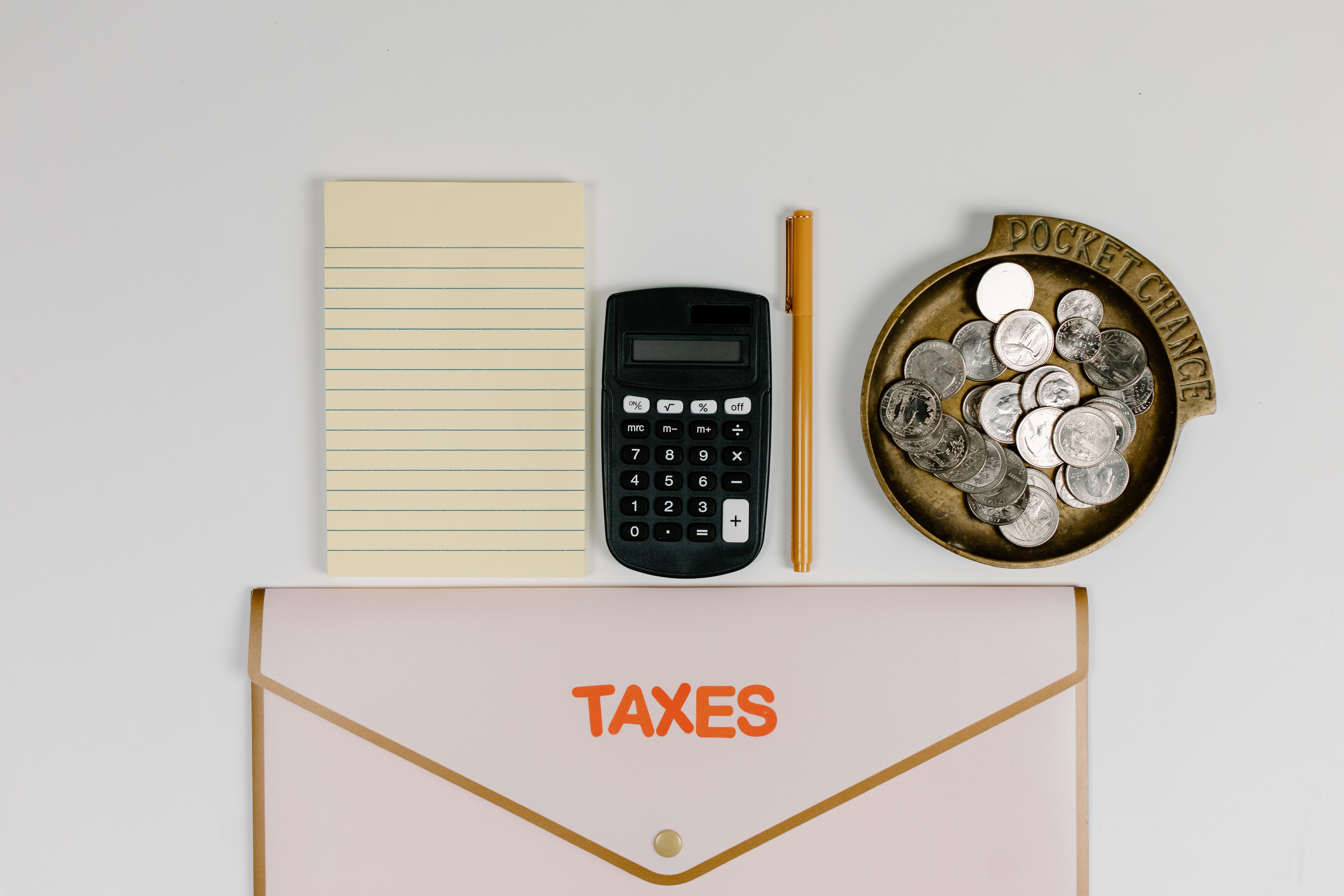 A flat lay of tax preparation essentials calculator, coins, notepad, pen, and folder.