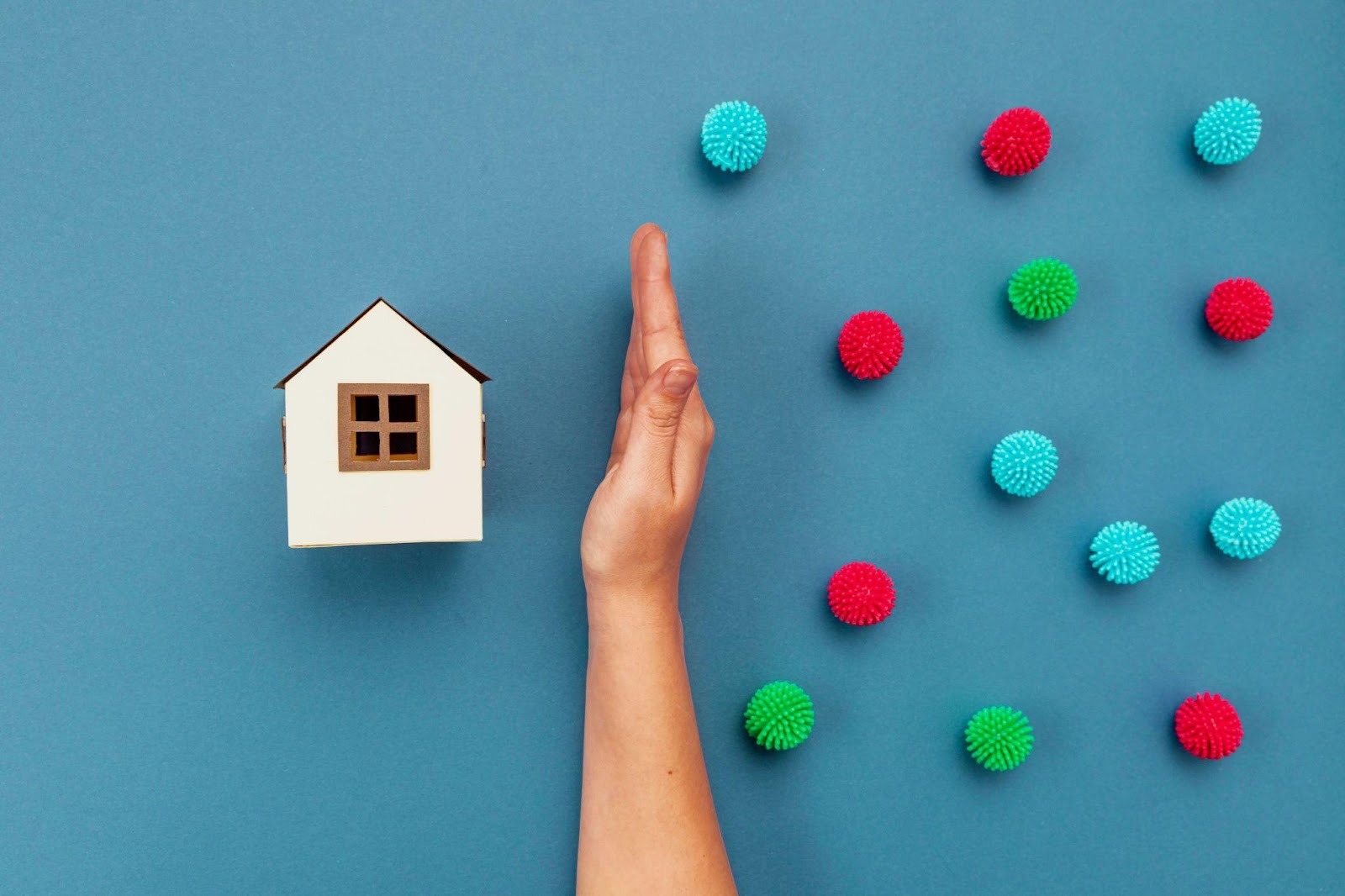 A hand shielding a small paper house from colorful spiky balls on a blue background.