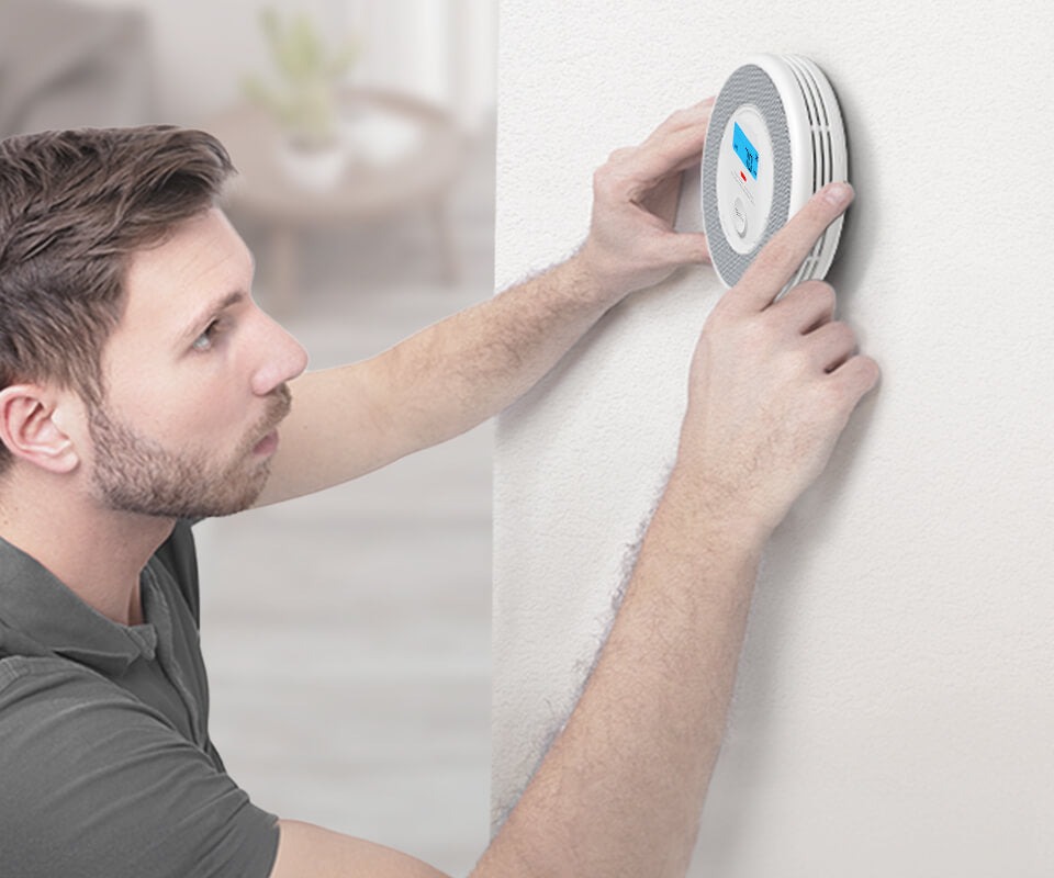 A man installing or testing a round white smoke and carbon monoxide detector on a plain wall.