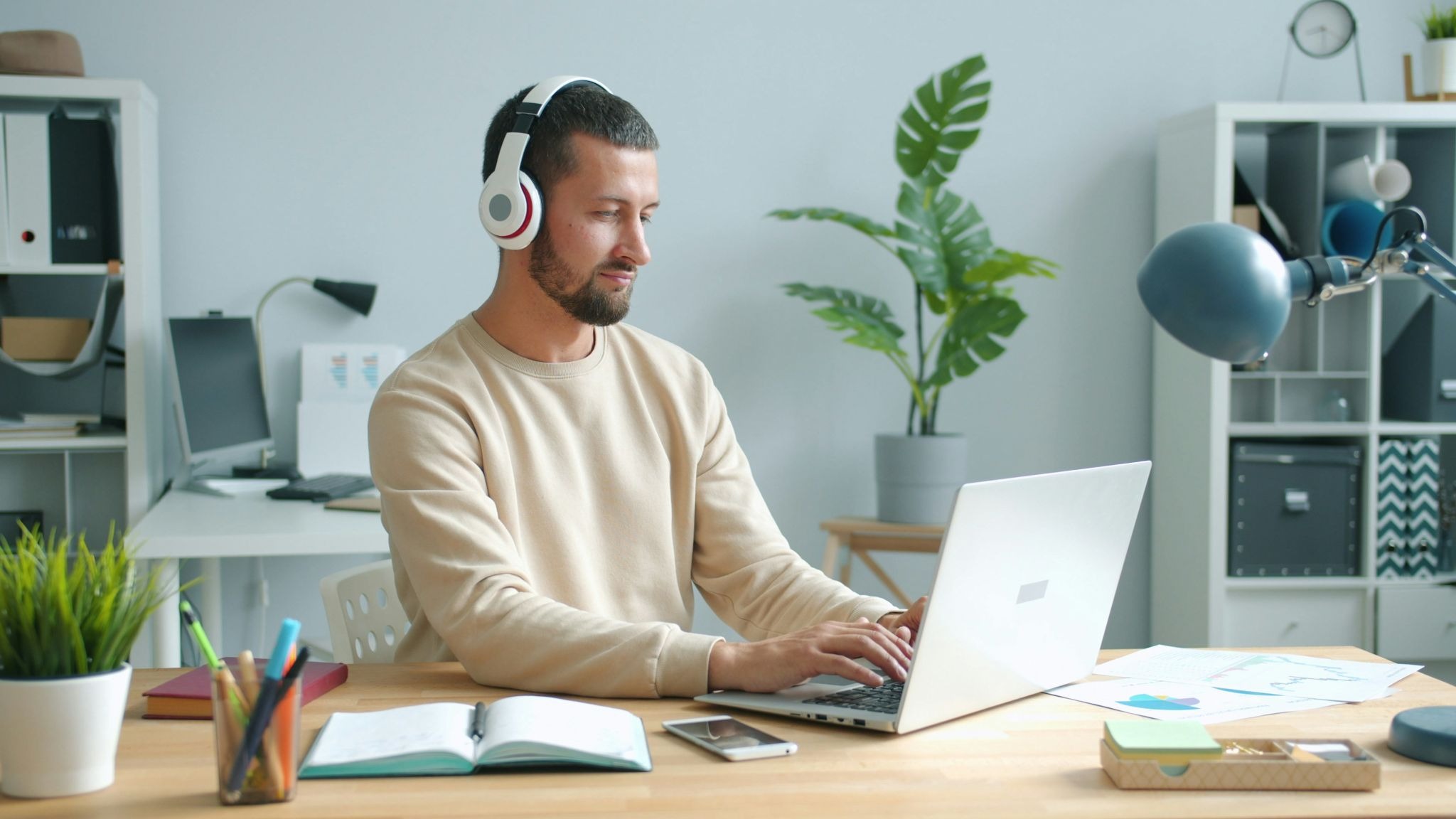A man with a beard wearing white headphones sitting at a desk and typing on a laptop in a bright home office.