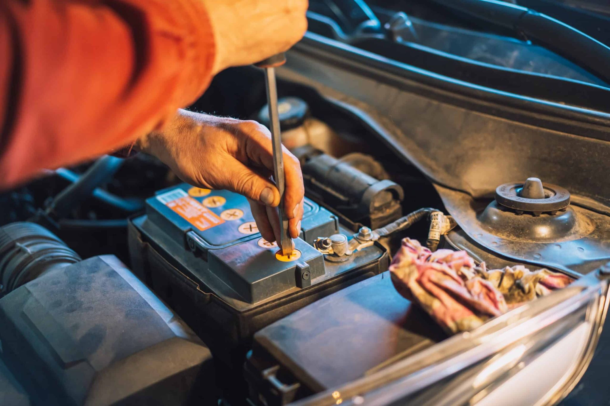 A mechanic's hands using a screwdriver to work on a car battery under the hood of a vehicle.