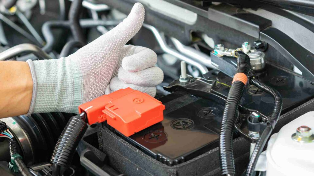 A person wearing a grey textured glove giving a thumbs-up next to a car battery with its red terminal cover.