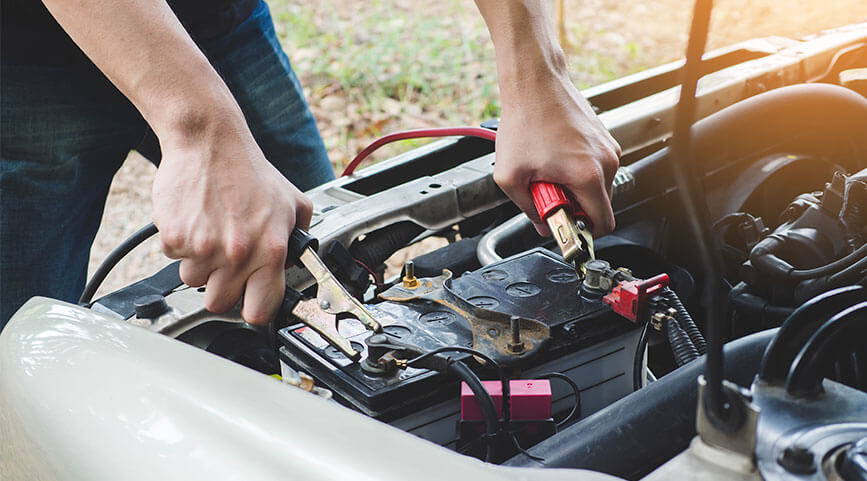 A person&rsquo;s hands using jumper cables to jump-start a car battery under an open hood.