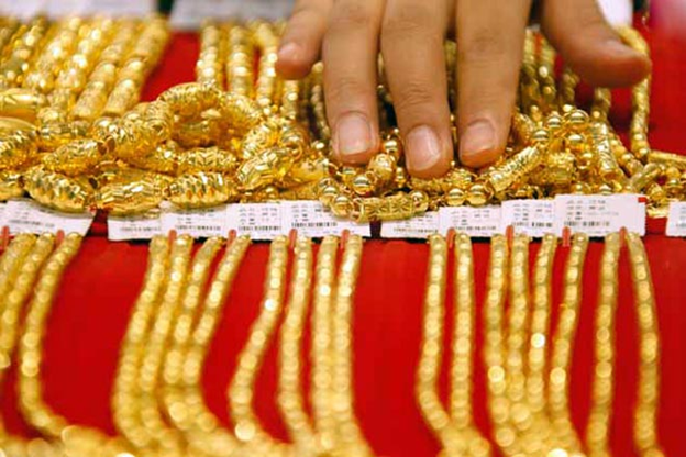 A person's hand is shown over many rows of gold bracelets and necklaces displayed on a red surface.
