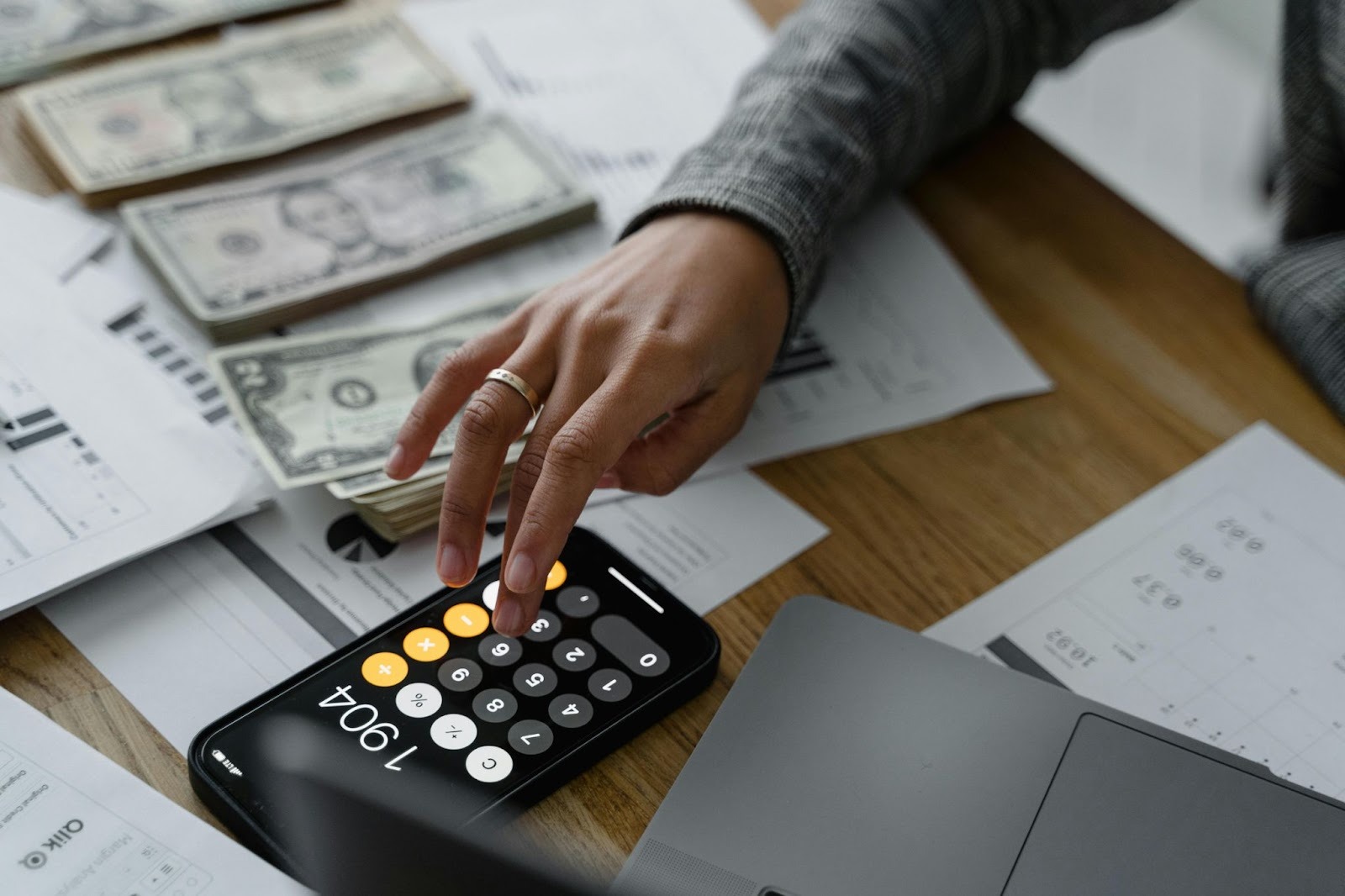 A person's hand using a calculator app on a smartphone next to stacks of US dollar bills and financial documents on a wooden desk.