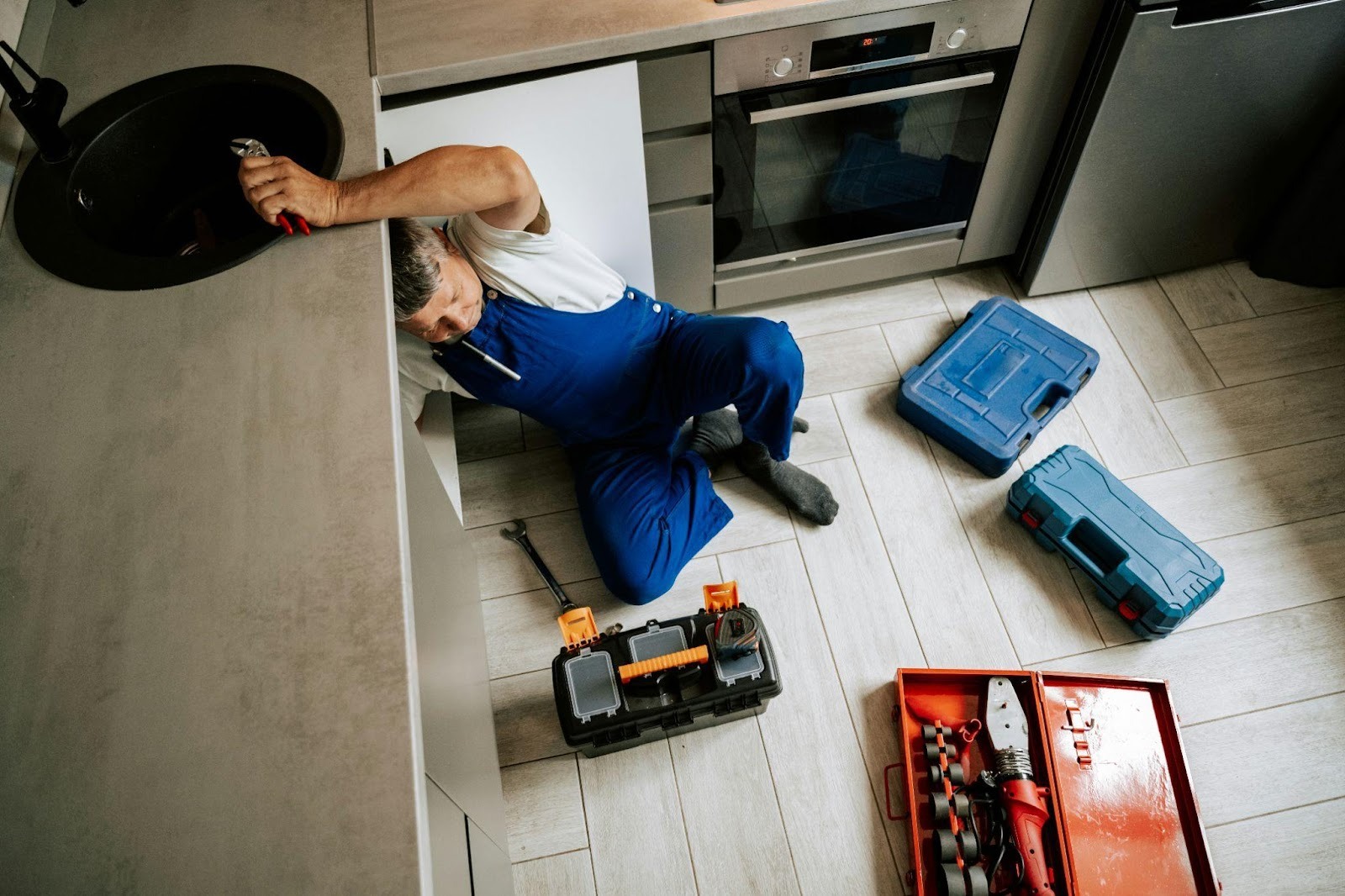 A plumber in blue overalls lying on the floor while repairing a kitchen sink, surrounded by various open toolboxes.