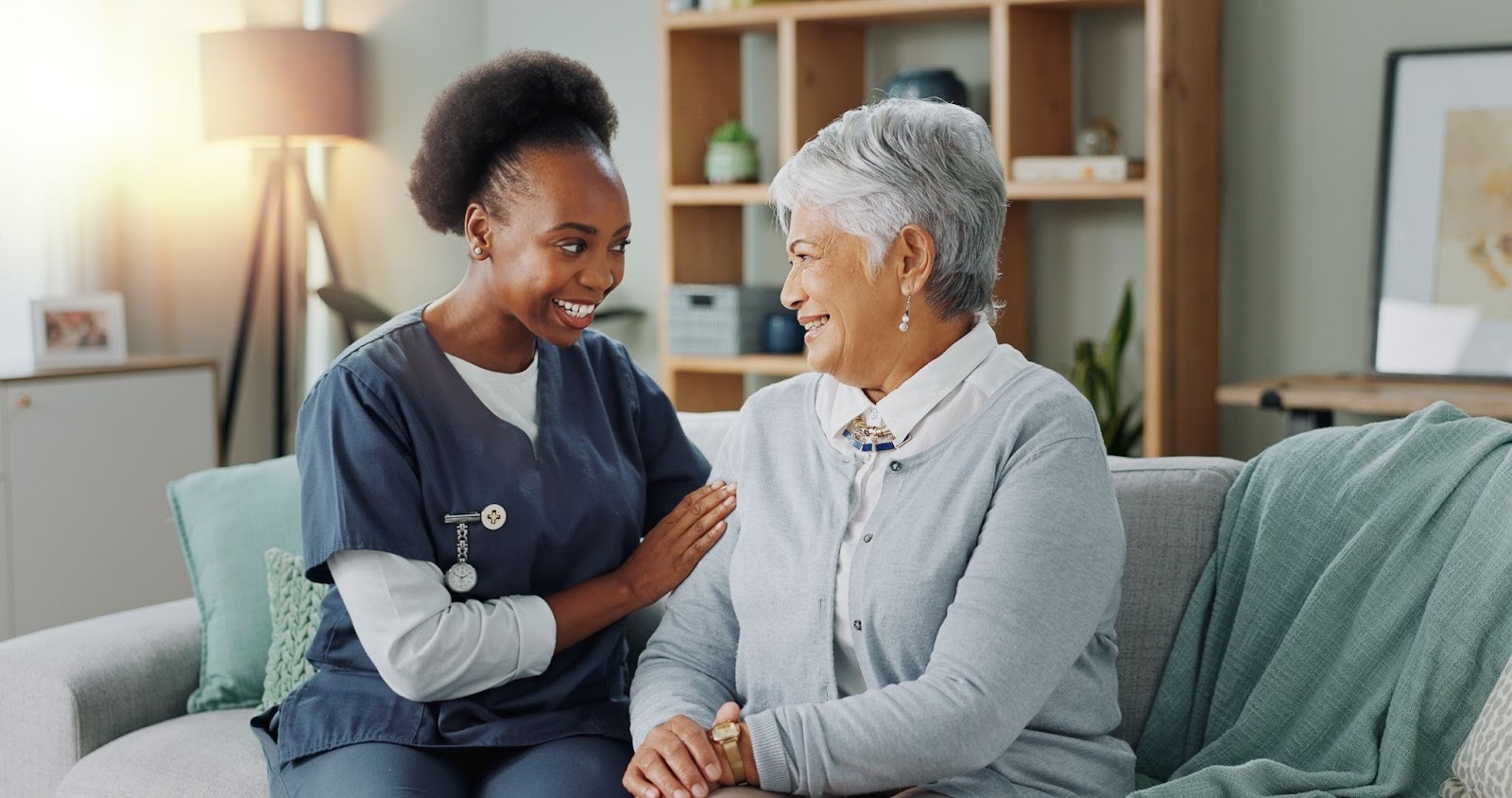 A smiling home healthcare worker in blue scrubs sitting on a couch and comfortingly placing a hand on the arm of an elderly woman.