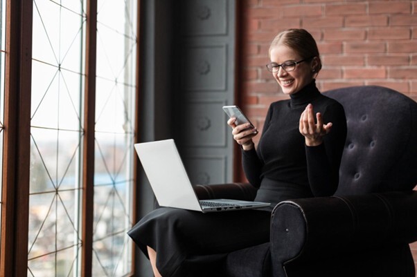 A smiling woman in a black turtleneck and glasses sitting in a dark armchair, holding a smartphone and looking at a laptop on her lap by a large window.