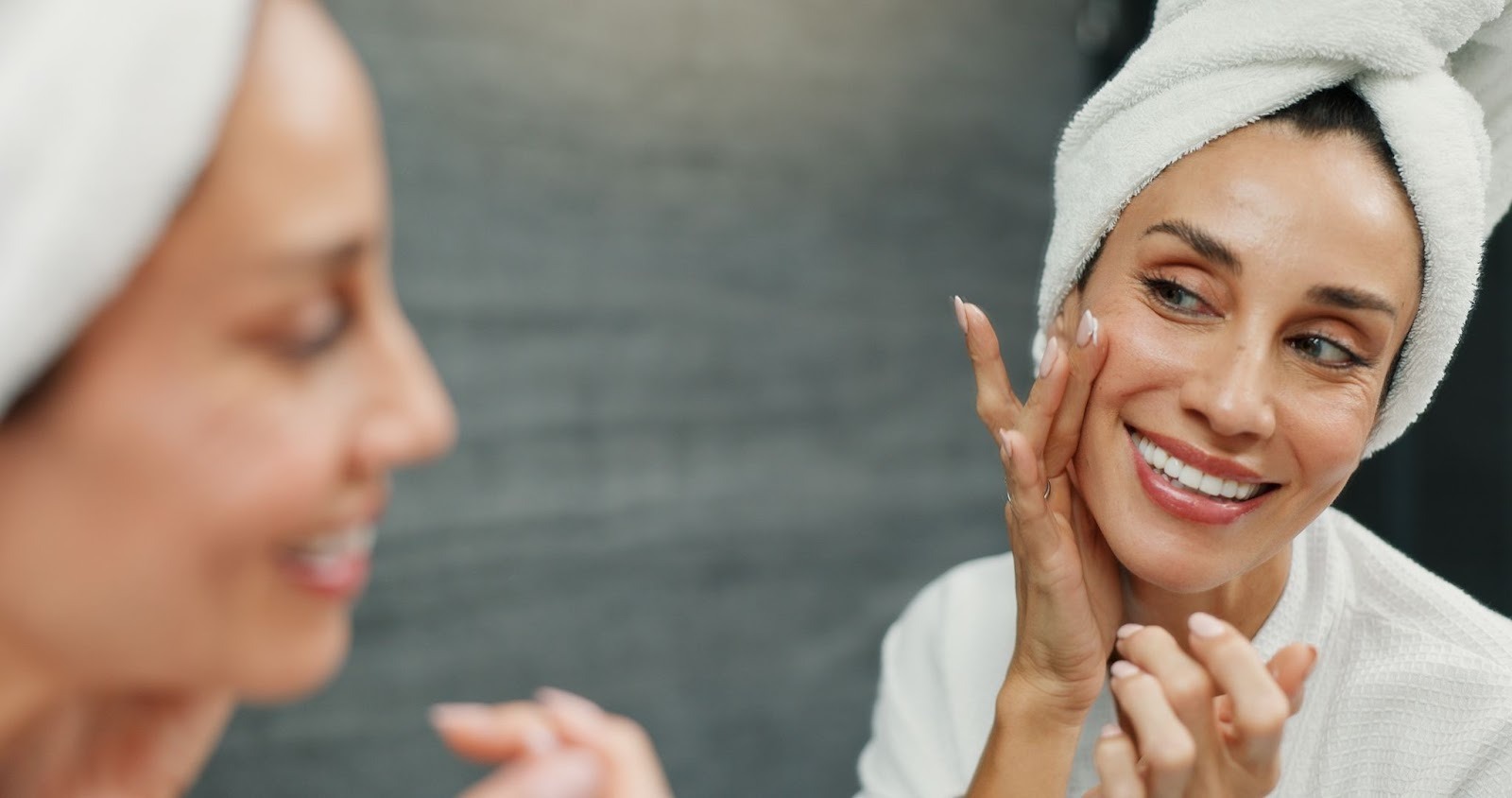 A smiling woman in a towel is applying cream to her cheek while looking at her reflection.