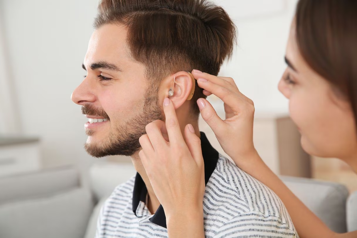 A woman gently adjusting a hearing aid in a man's ear as he smiles.