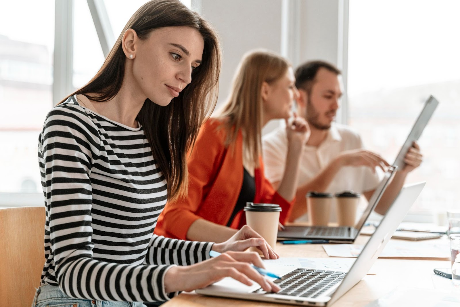A woman in a striped shirt working on a laptop while two colleagues talk and use a tablet in the background.