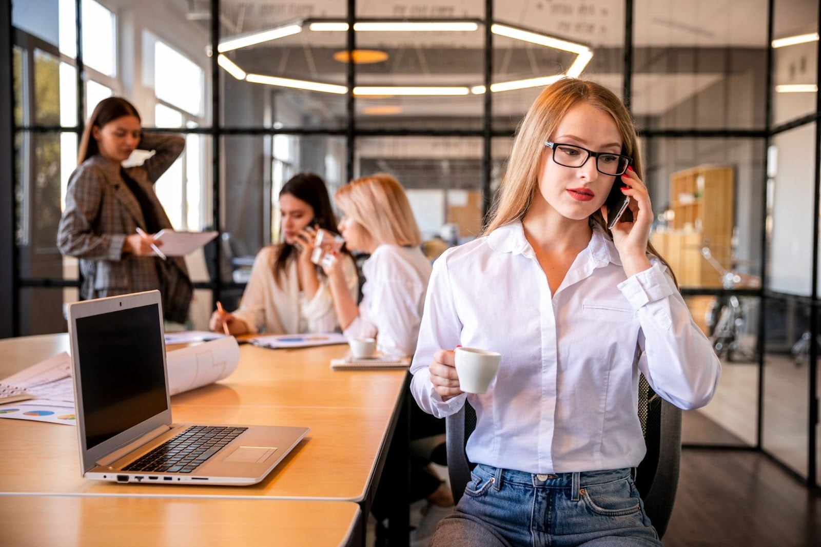 A woman in a white shirt and glasses talking on a smartphone and holding a coffee cup in a modern office.