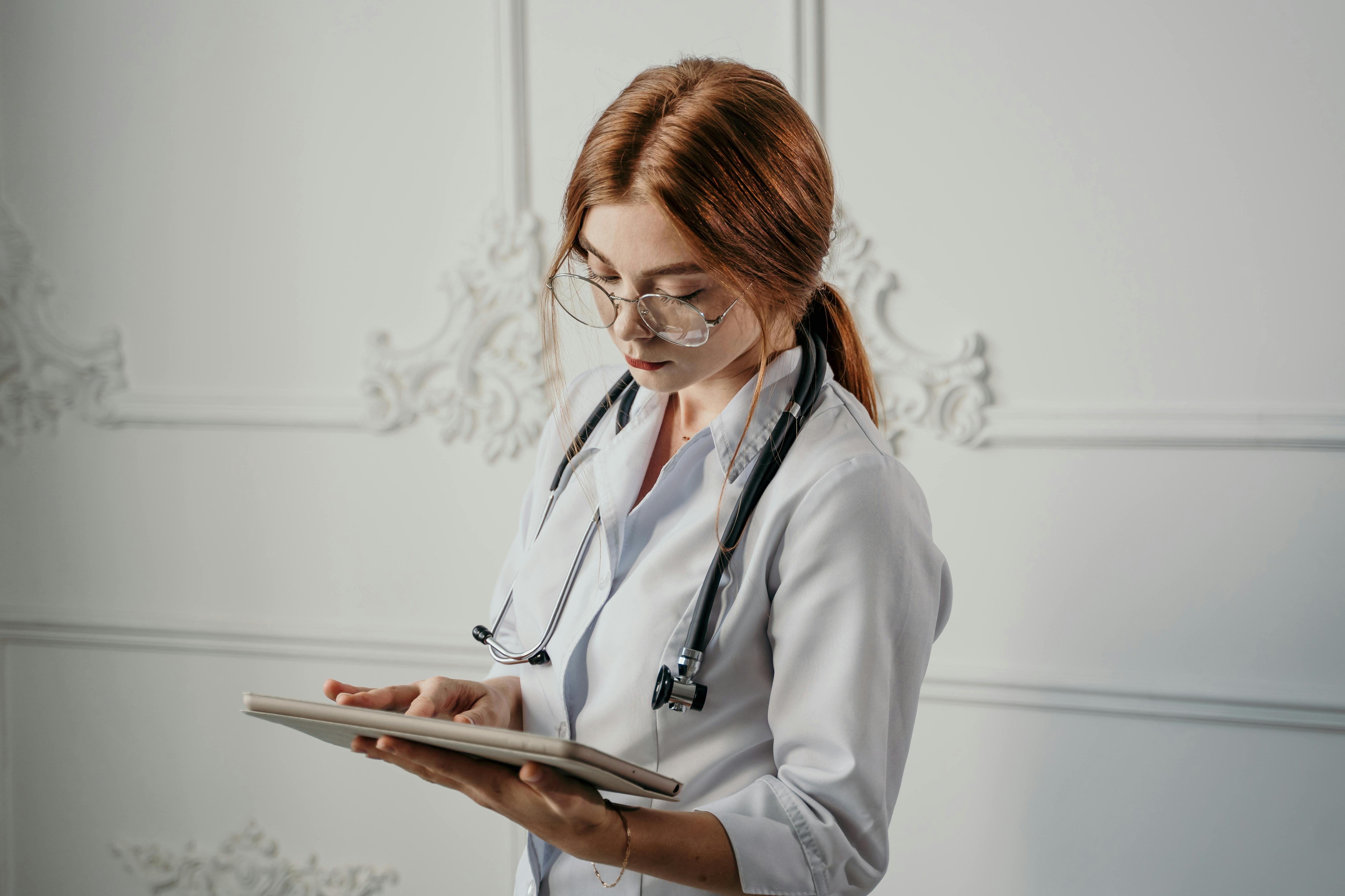 A young doctor or medical professional in a white coat with a stethoscope around her neck, focused on using a digital tablet in her hands.