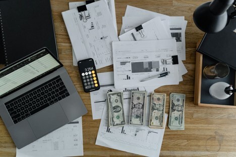 Laptop, printed financial reports, calculator app on a smartphone, and various U.S. dollar bills spread across a wooden desk.
