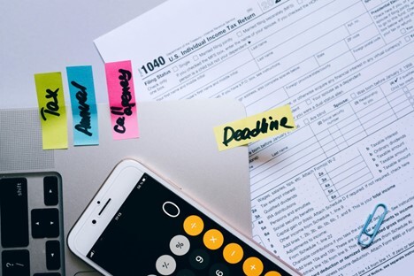 Sticky notes labeled “Tax,” “Annual,” “Challenge,” and “Deadline” on a laptop next to a smartphone calculator and a U.S. tax form.