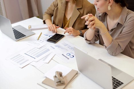Two women discussing financial documents at a desk with laptops, a calculator, and printed charts.