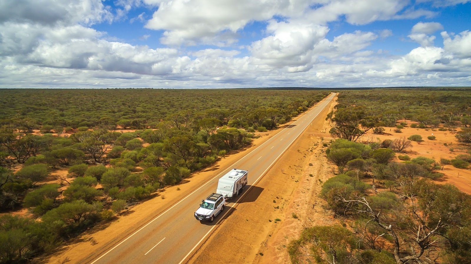 Aerial view of a white car towing a caravan down a long, straight road through the arid Australian outback.