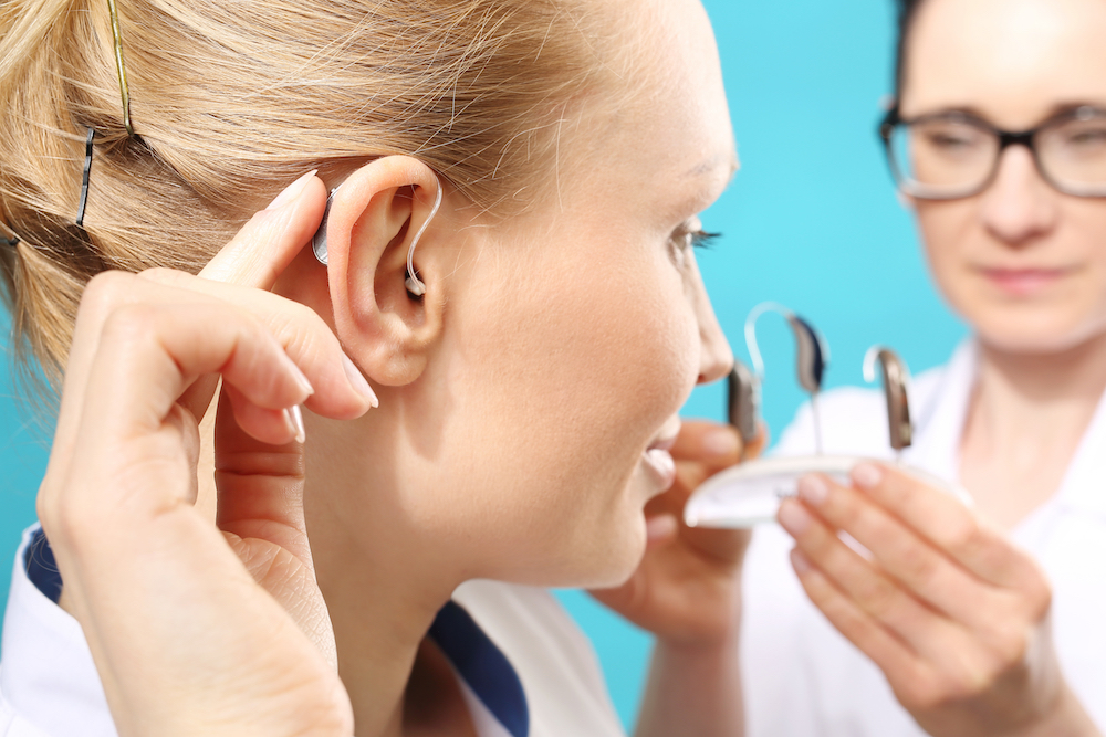 An audiologist in a white coat showing a woman different hearing aid models while she wears one behind her ear.