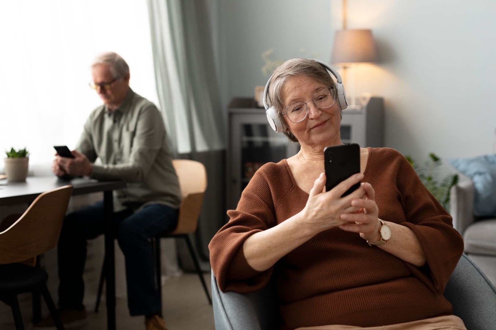 An elderly woman wearing headphones and glasses smiling while using her smartphone, with an elderly man using his phone in the background.