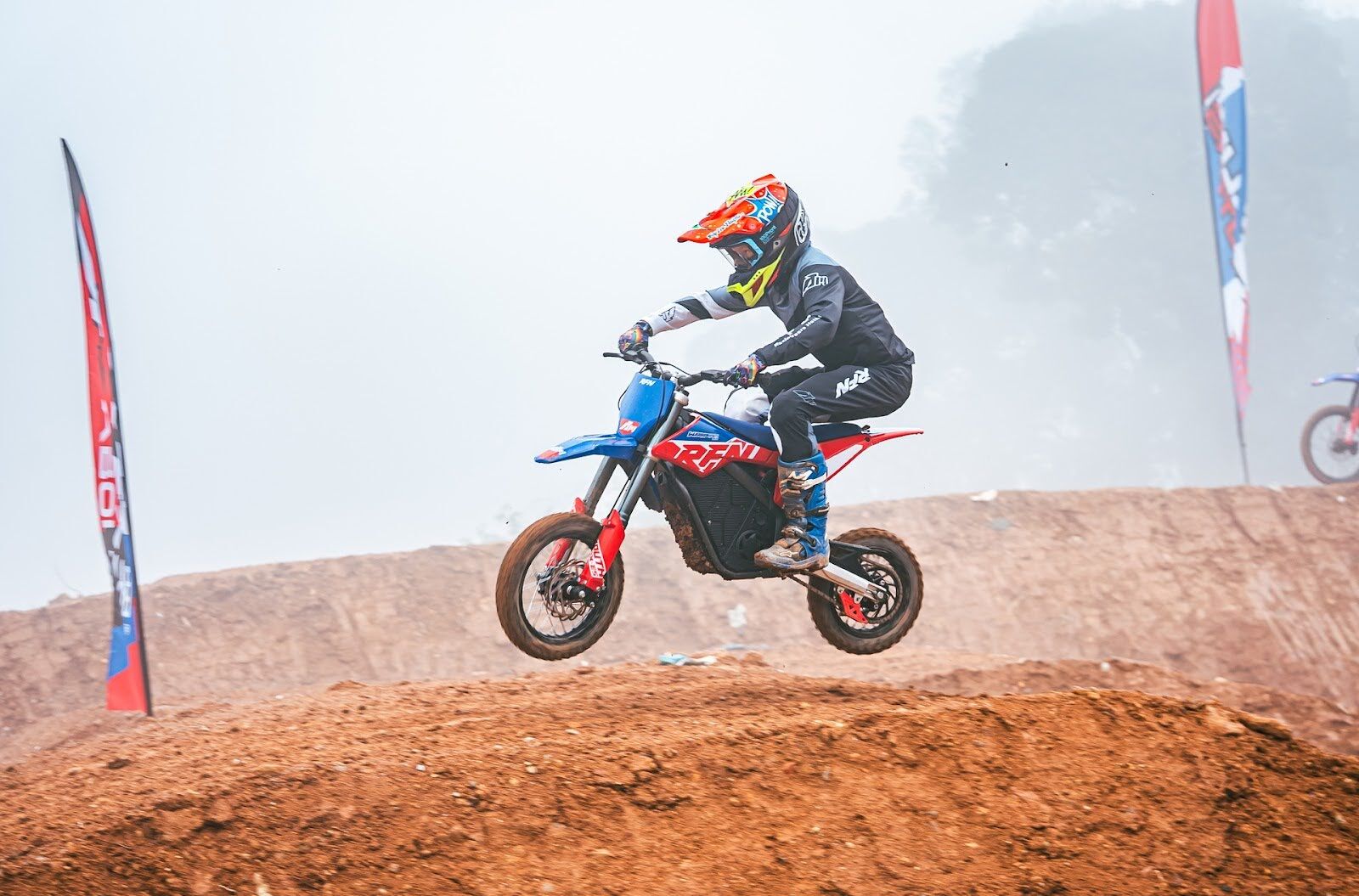 A motocross rider wearing full gear, including a red and blue helmet, jumps a dirt mound on a red and blue electric dirt bike, with flags and a misty backdrop.
