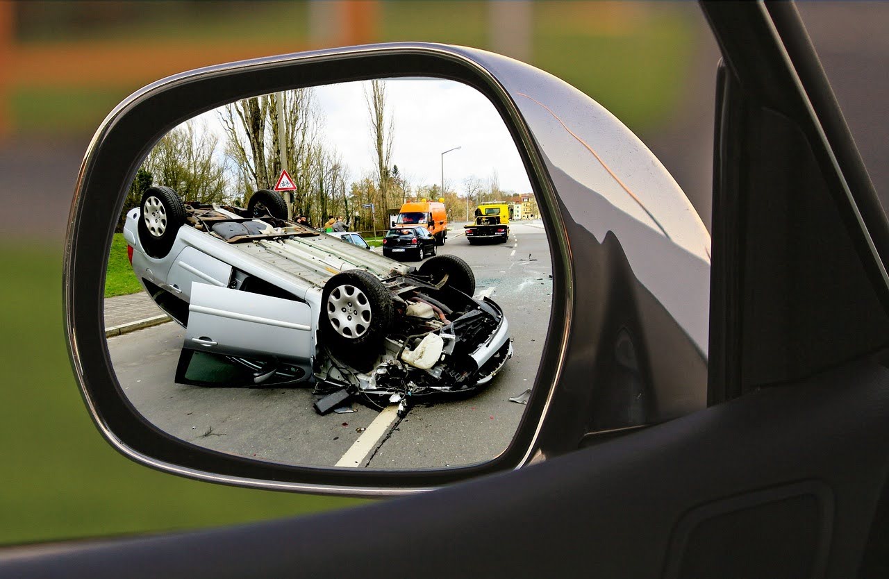 A car's side mirror reflects a flipped silver car on a road, the aftermath of an accident, with other vehicles nearby.