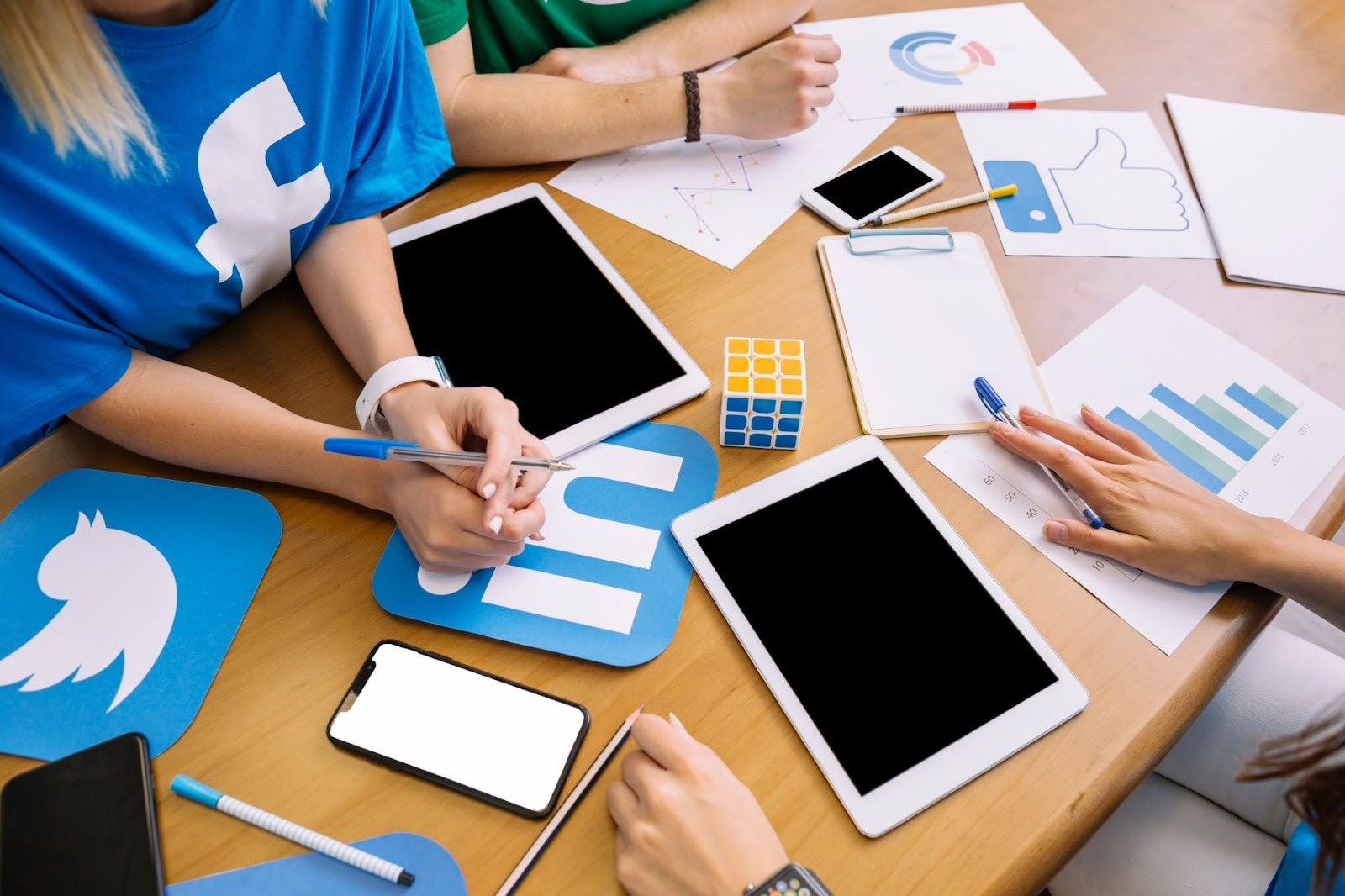 Close-up of a team meeting around a table with social media logos, tablets, phones, and marketing graphs.