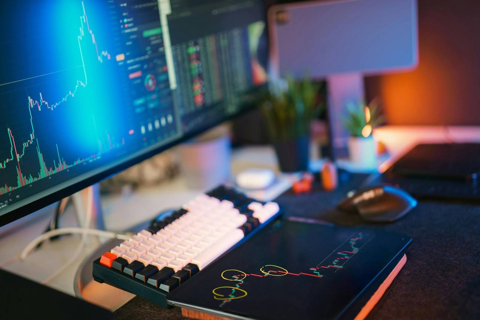 Computer desk setup with a monitor showing stock charts, a mechanical keyboard, and a tablet displaying highlighted trading data.