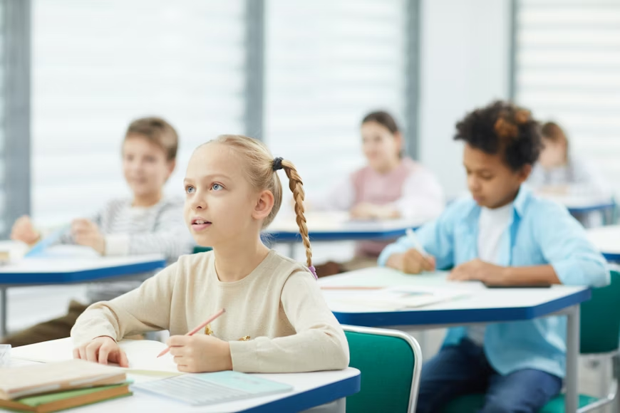 Curious little girl with blond hair sitting at school desk listening to her teacher