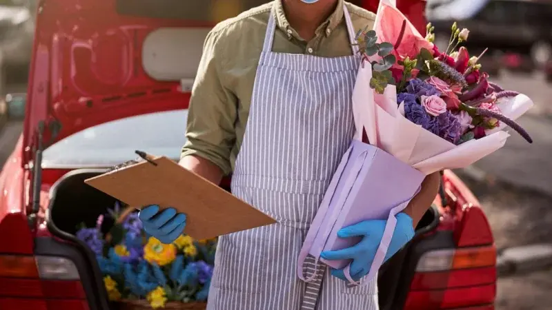 A person wearing gloves and an apron, holding a bouquet of flowers and a clipboard next to a red car trunk.